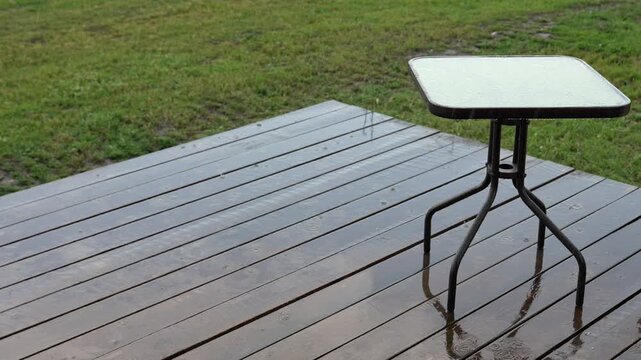 Summer rain falling on a wet wooden deck and an empty outdoor table with a green lawn in the background, creating a calm and relaxing scene with visible raindrops and reflections