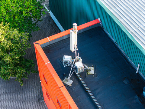 Aerial view of a cellular antenna base station mounted on a flat rooftop with an orange facade and green metal roof section in Riga, Latvia.