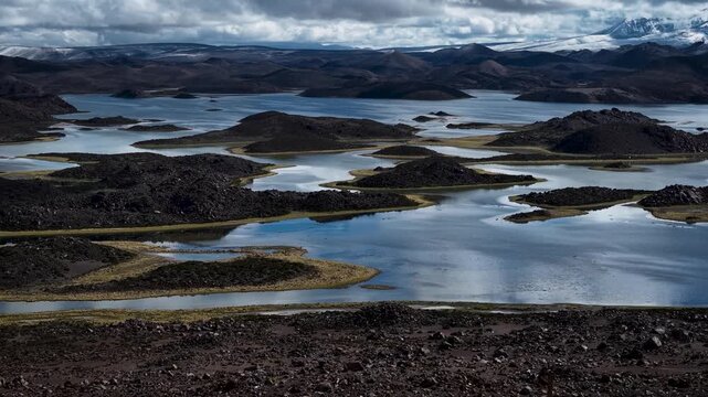 Elevated oblique view reveals expansive volcanic lake system with dark lava islets, sinuous shallow channels, cool reflections, rugged peninsulas, and quiet remote scenery under overcast light
