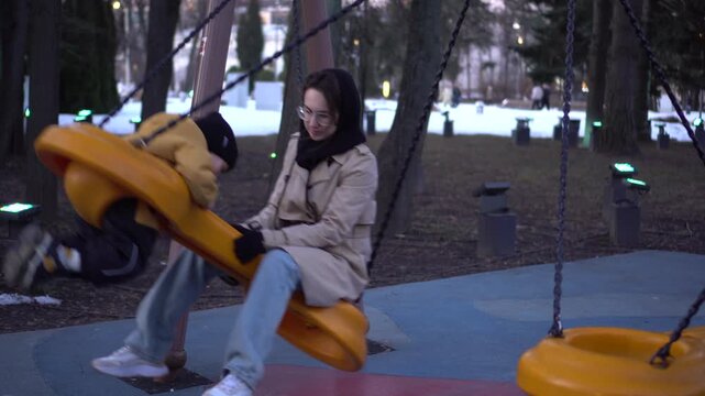 Woman pushing a child on a yellow swing in a winter park at dusk