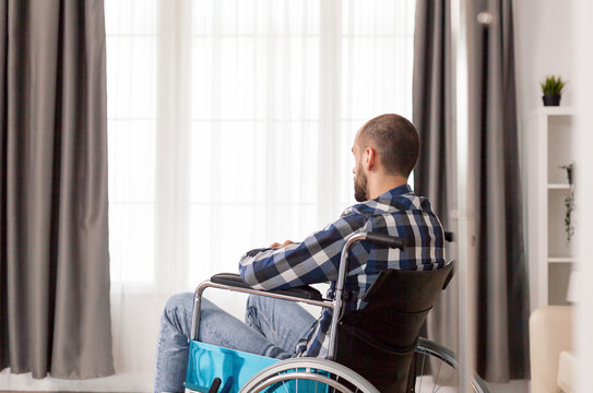 Amputee man seated in wheelchair in medical office sitting in wheelchair in the middle of medical office. Serious sad caucasian man wearing casual clothes and look at large panoramic view in bright.