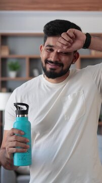 A smiling man of Indian descent wipes sweat from his forehead while holding a blue water bottle after a home workout