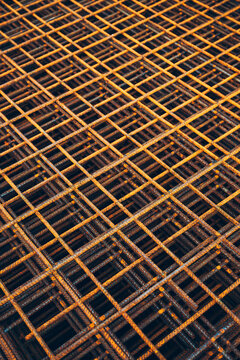 Rusted steel rebar grid stacked at a construction site, creating a repeating geometric mesh pattern. Strong industrial metal texture and warm orange rust tones.