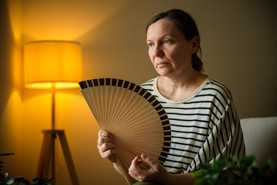 Woman using a handheld fan to cool herself during an indoor summer heatwave. Climate change, energy conservation and household comfort concept.