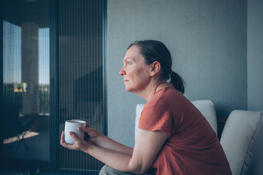 Middle-aged woman sits on a balcony holding a coffee cup and gazes thoughtfully into the distance. Quiet, reflective morning moment.
