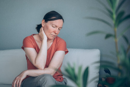 Mature woman suffering from severe neck pain while sitting on balcony sofa, seen through oleander leaves. Musculoskeletal discomfort and health problem concept.