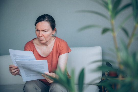 Anxious middle-aged woman reviewing debt notice and paperwork while sitting outdoors. Framed by plant foliage, conveying financial problems, budgeting challenges, and household expenses.