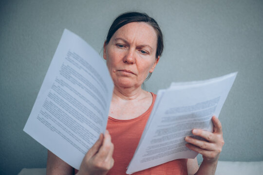 Worried mature woman reading debt notice and financial documents while sitting on balcony sofa. Concerned expression reflects personal finance problems, unpaid bills and economic stress.