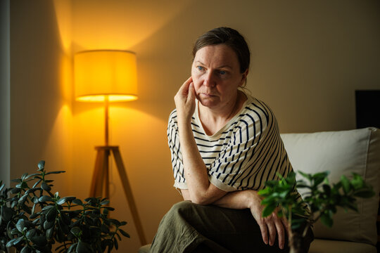 Middle-aged woman with a serious expression deep in thought in a comfortable living room. Contemplation, life choices and mental wellbeing theme.