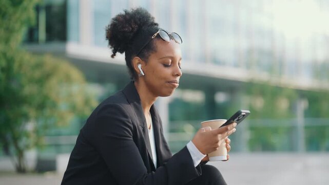 Happy townswoman calling by mobile, listening sound by earphones, drink coffee. Good morning in metropolis, businesswoman discussing working asks by phone outdoors, modern technology for work and rest
