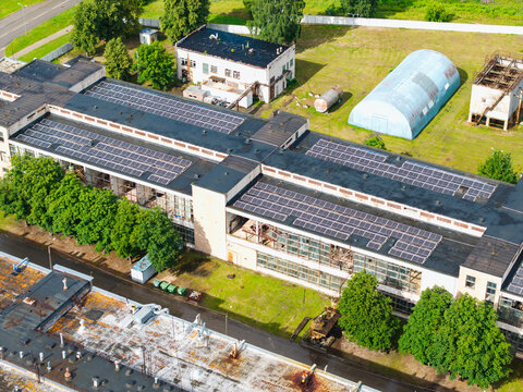 Aerial view of an industrial building featuring a large rooftop solar panel array surrounded by green trees and paved grounds in Olaine, Olaine Municipality, Latvia.
