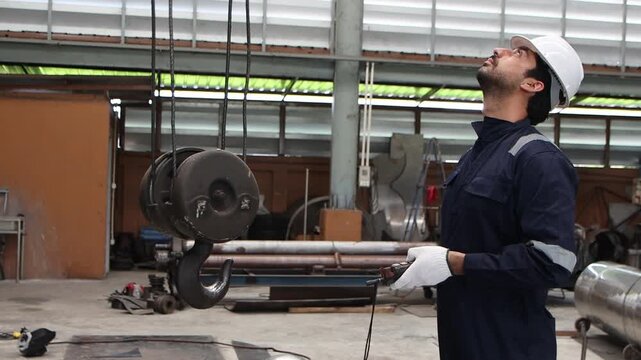 Industrial engineer supervisor foreman in safety suit and white helmet working to control machinery to lift a large iron hook up at manufacturing plant. Worker operates a lift machine in metal factory