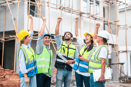 A group of happy Asian construction workers celebrating success by raising their hands in the air at a construction site. Teamwork, collaboration, safety, achievement, and positive workplace culture i