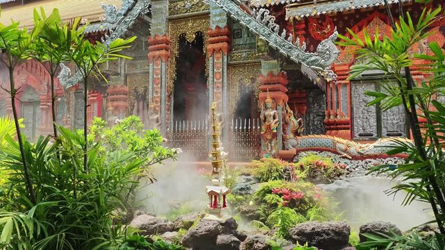 A mist machine creates an ethereal fog through a lush tropical garden at an ornate Thai Buddhist temple, with red, gold, and silver-toned facades, naga serpent roof carvings, and a small spirit shrine
