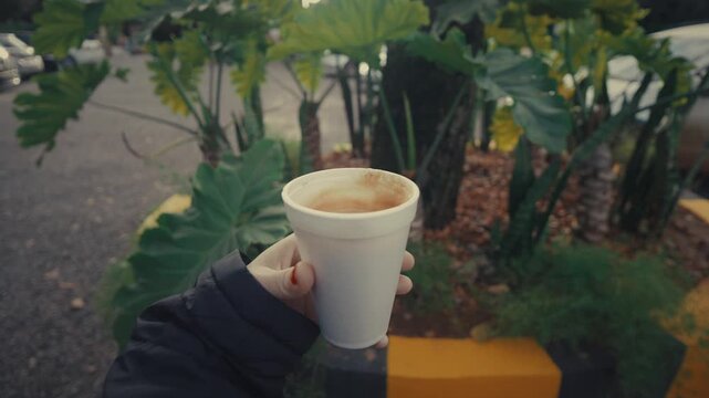 Hand holds a white coffee cup. Foam appears on the surface. Tall green plants border the street. The person wears a dark jacket. This moment feels calm. Perfect for coffee or outdoor scenes