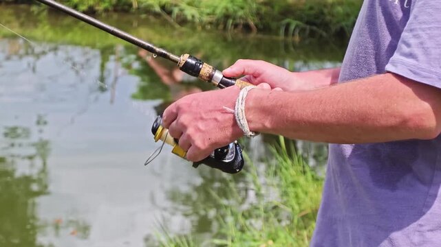 In a fixed, steady shot, a man's hands grip a spinning fishing rod and reel beside a calm green pond, with reflections of grass and trees visible in the water. Filmed at 120fps and conformed to 30fps