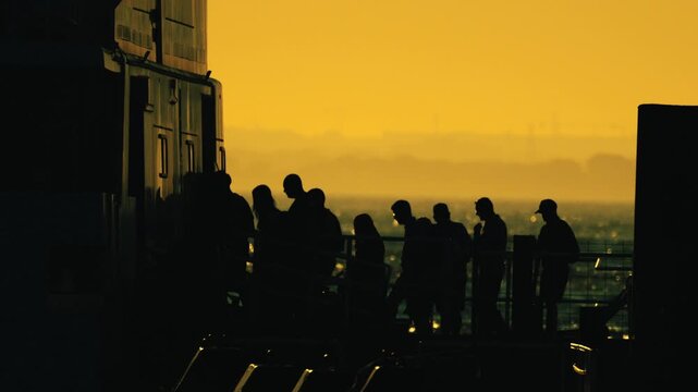 Silhouetted group of people boarding a ferry at sunset, with warm golden hues illuminating the scene and the ocean visible in the background