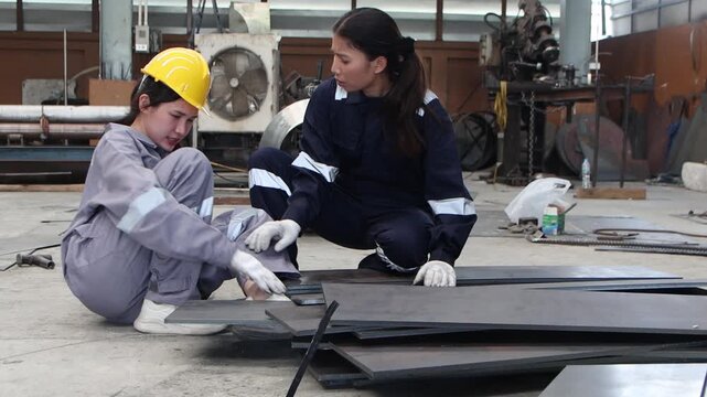 Two female engineer workers in industrial safety suit and helmet try to lift up heavy metal plate before it fell on her foot at manufacturing factory. Staff has accident and her colleague try to help.