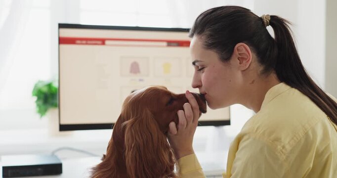 Dog owner woman and spaniel browsing online. At home by a computer monitor, she kisses her pet as a retail site shows product thumbnails. Cozy daylight. Theme is pet friendly online shopping.