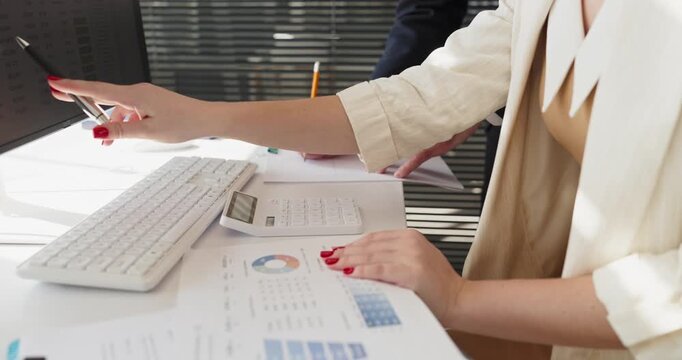 Coworkers conduct finance analysis with calculator and charts. Hands point at computer screen reviewing reporting, data and charts beside a keyboard. Clear office business teamwork concept.