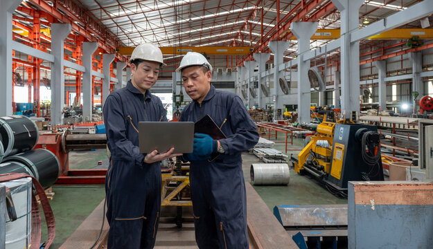 Two workers in safety helmet and mechanic coverall dust protection suit look at laptop computer, standing in a factory. They check information and discuss their task.