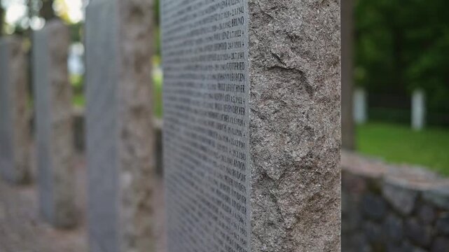 Engraved stone monuments stand in a garden, surrounded by green grass and trees. The stones display detailed text and create a reflective environment for visitors