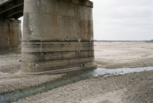 Concrete bridge pillar with old water level marks rises from a nearly dry riverbed with only a small stream remaining.