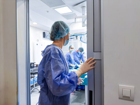 Female surgeon in blue scrubs and protective mask standing at the doorway of a sterile operating room, observing a surgical team performing a procedure. A view into a professional hospital theater.