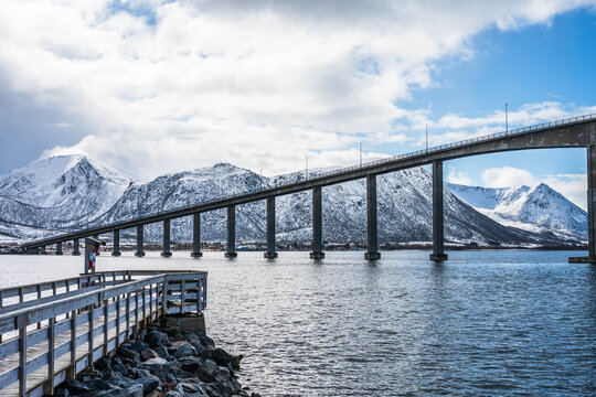 Risoyhamn on Andoya Island in Norway shows a bridge over calm waters with mountains in the background and a path along the shore during a cloudy day
