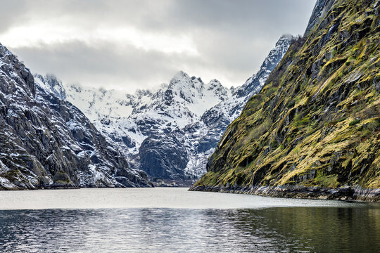 Trollfjord in Norway shows steep cliffs and snowy peaks surrounding the water on a cloudy day with reflections on calm water