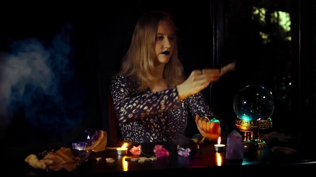 Woman psychic performs a smoke purification ritual over a crystal ball with candles amethyst and runes in a dark mystical space showing focused esoteric practice