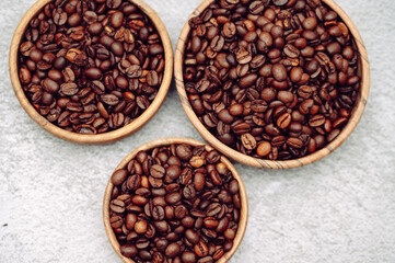 Fototapeta premium Three round wooden bowls filled with roasted brown coffee beans on a grey concrete background.Aromatic dark roast coffee beans arranged in circular wood plates, top view copy space.
