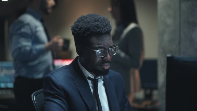 African American male broker wearing glasses looking at computer screen in dark trading office. Trader reacting to financial chart. Businessman discussing stock market data. Team arguing behind him.