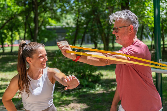 Senior man performing a resistance band chest fly exercise under the guidance of a fitness coach to improve upper body strength, shoulder stability, posture, and functional movement outdoors.