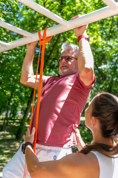 Senior man performing an assisted pull-up with resistance bands under the supervision of a fitness coach to improve upper body strength, grip, mobility, and functional fitness.