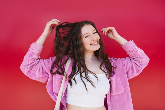 Smiling portrait of woman winking and touching hair against red background