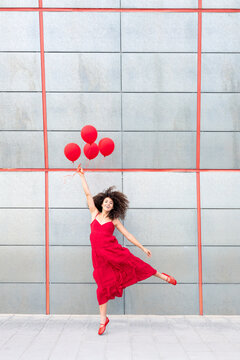 Smiling woman in red dress jumping with balloon by grey wall