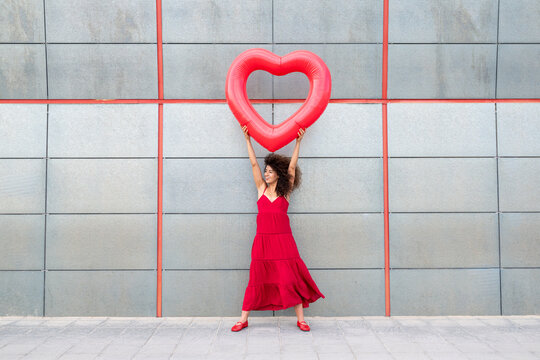 Smiling woman in a red dress holding a big red heart against a grey wall