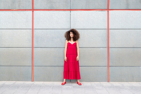 Model in red dress with curly hair standing against grey wall