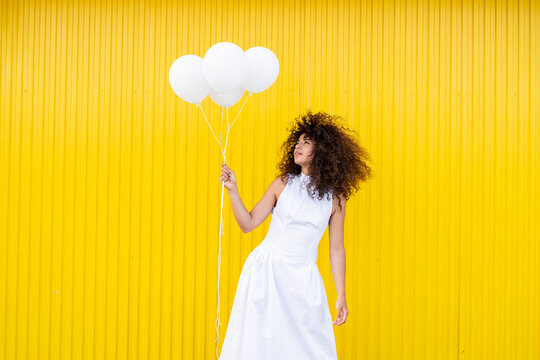 Woman in white dress holds white balloon against yellow background