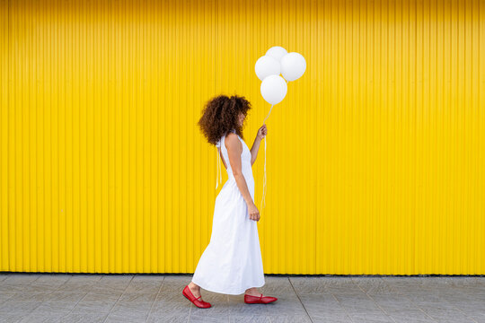 A woman in a white dress walks with a balloon by a yellow wall