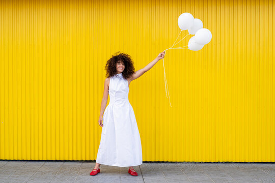 Smiling woman in white dress holds white balloon against yellow wall