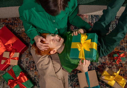 Red-haired boy smiling with mother and holding gift at home on carpet