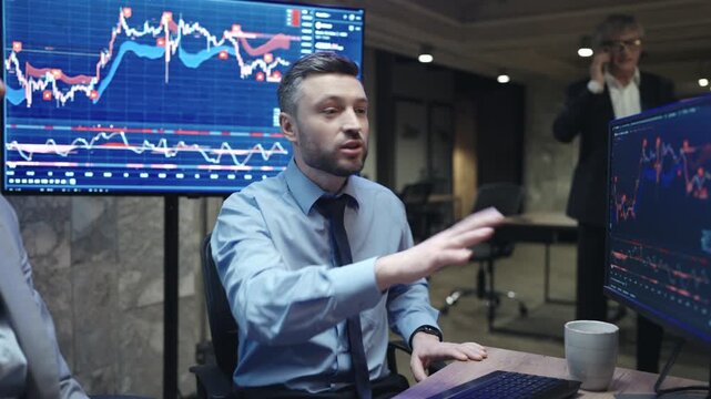 Male broker gesturing toward financial chart monitor while explaining data to female colleague. Trading team discussing stock market strategy in dark office. Senior manager calling at night.