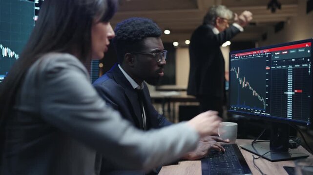 African American male broker typing on keyboard while watching falling financial chart. Female colleague sitting beside him in dark trading office. Senior manager standing behind team at night.