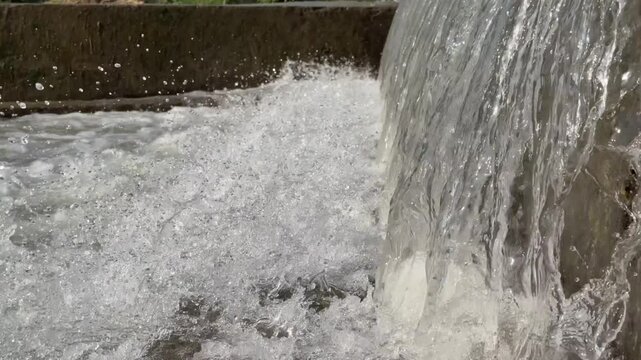 Artificial concrete check dam directs water into churning, frothy river stream lined with green trees.