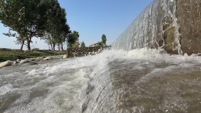 Artificial concrete check dam directs water into churning, frothy river stream lined with green trees.