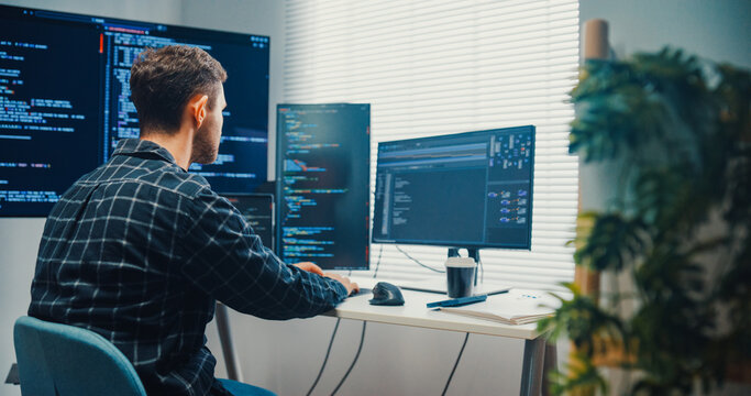 Young Caucasian male programmer writes code using multiple screens in home office workspace. AI development, cybersecurity, network database system, software engineering, remote tech startup concept.