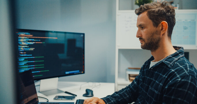 Young Caucasian male programmer writes code using multiple screens in home office workspace. AI development, cybersecurity, network database system, software engineering, remote tech startup concept.