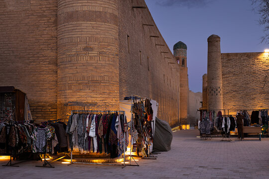 Textile market stalls along the fortress walls of Ichan Kala in Khiva at dusk. Warm ground lights illuminate ancient brick towers and colorful scarves displayed on racks. Ideal for travel, tourism.